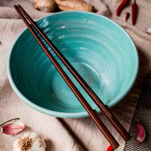 Turquoise bowl with chopsticks from above on a cream tablecloth with garlic, chilli and ginger scattered around.