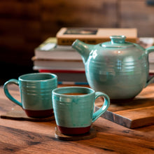 Load image into Gallery viewer, 2 turquoise mugs and a turquoise teapot sitting on a wooden table with books in the background.