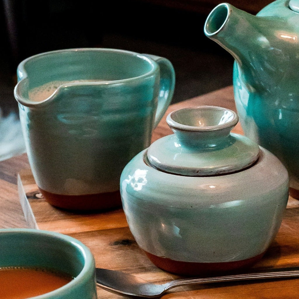 Turquoise milk jug and sugar bowl, teapot in the corner, teaspoon at the front on a wooden table.