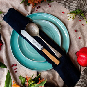 A turquoise side plate sitting on a turquoise dinner plate with a navy napkin, knife and spoon ontop. On a beige tablecloth with flowers and pomegranate scattered around.