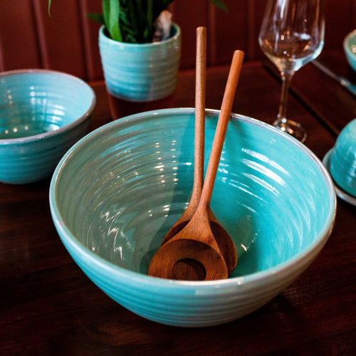 Large turquoise bowl with 2 wooden serving spoons inside. A turquoise bowl, vase and wine glass in the background.