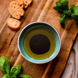 Small turquoise bowl with olive oil and balsamic on a chopping board with mini bread slices and basil.