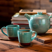 Load image into Gallery viewer, 2 turquoise mugs and a turquoise teapot sitting on a wooden table with books in the background.
