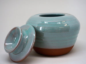 Turquoise jar with a lid leaning against iton a white background.